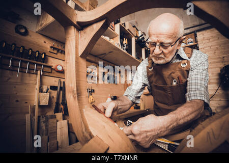 An old carpenter takes measurements at a chair to be restored, a workshop with tools in the background. Stock Photo