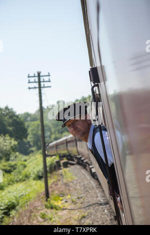 Close up portrait of smartly-dressed train guard in peaked cap, on board vintage UK steam train, leaning out of railway carriage window looking out. Stock Photo