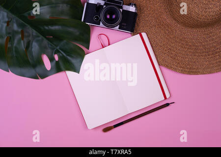 Women's traveler accessories: straw hat, tropical leaf monstera, retro camera, notebook with copy space, paint brush on pink background. Summer concept of travel background Stock Photo