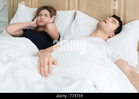 Annoyed Asian wife blocking her ears from noise of husband snoring in bedroom at home. Young couple have problem with man's snoring. Stock Photo