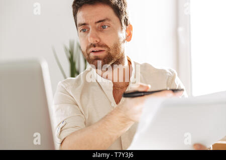 Photo of concentrated office worker 30s wearing white shirt using laptop and paper documents while sitting at table in modern workplace Stock Photo