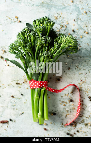 high angle view of a bunch of broccolini, tied with a red ribbon patterned with white dots, placed on a white stone surface Stock Photo