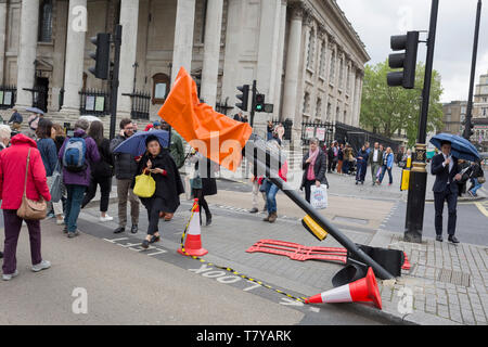 During seasonal spring rain, pedestrians cross Trafalgar Square and around the leaning traffic light post, damaged after a recent vehicle crash, on 9th May 2019, in London, England. Stock Photo