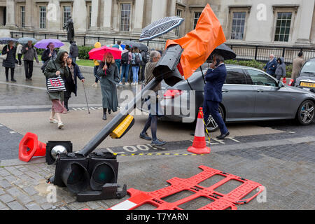 During seasonal spring rain, pedestrians cross Trafalgar Square and around the leaning traffic light post, damaged after a recent vehicle crash, on 9th May 2019, in London, England. Stock Photo