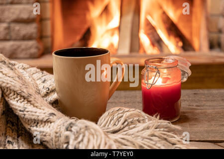 Big mug with hot tea, and a candle , wool scarf, near cozy fireplace, in country house, hygge, home sweet home. Stock Photo