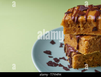 Stack of three sweet potato blondies with thin lines of chocolate and caramel icing running down at the sides, chocolate chips on teal plate, green ba Stock Photo