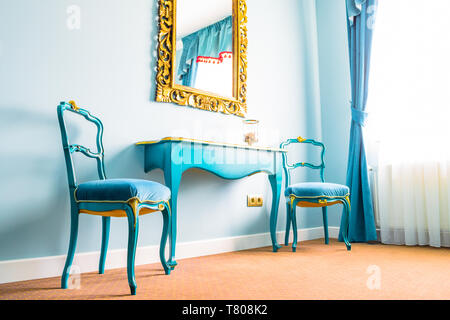 Two blue vintage chairs and a wooden table, next to a wall with a mirror, decorated with yellow floral swirls on its frame. Blue curtains on the side  Stock Photo