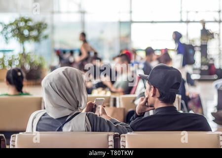 Modern muslim islamic asian couple using their smartphone apps while sitting and waiting for flight departure at international airport terminal Stock Photo