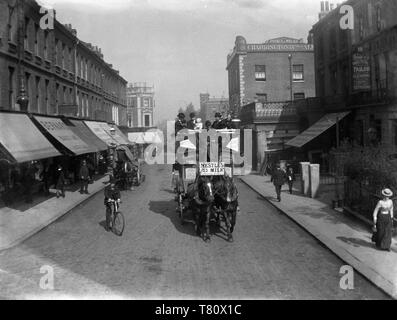 A Victorian London street scene of a horse drawn bus and other people going about their daily business, including a policeman, man on bike and smartly-dressed woman, with a back drop of canopied shops and The Prince of Wales Public House. Note the advert for Nestle's milk on the bus. Photo by Tony Henshaw Stock Photo