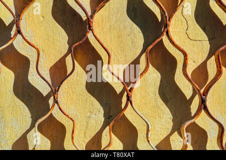 The texture of a yellow old plastered wall covered with cracks and closed metal grille. Stock Photo