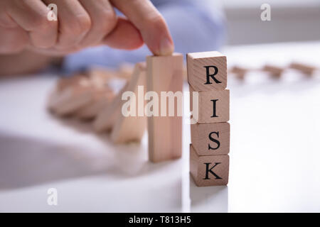 Businessperson's Finger Over Risk Wooden Blocks Stopping Dominos From Falling On Risk Word Desk Stock Photo