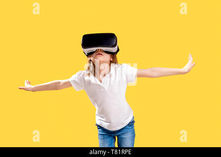 Girl 7 y.o in formal outfit wearing VR glasses putting hands out in excitement isolated on yellow background. Child using a gaming gadget for virtual reality. Virtual technology. Stock Photo
