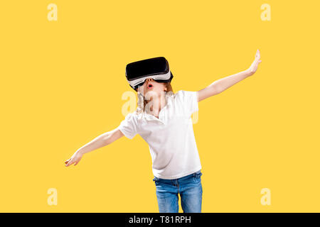 Girl 7 y.o in formal outfit wearing VR glasses putting hands out in excitement isolated on yellow background. Child using a gaming gadget for virtual reality. Virtual technology. Stock Photo
