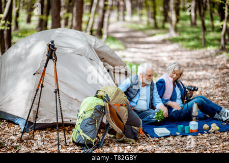 Senior couple sitting together at the campsite with tent and backpacks, enjoying nature with binoculars in the forest Stock Photo