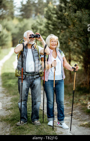 Happy senior couple standing together with binoculars, backpacks and trekking sticks while hiking in the forest. Concept of an active lifestyle on retirement