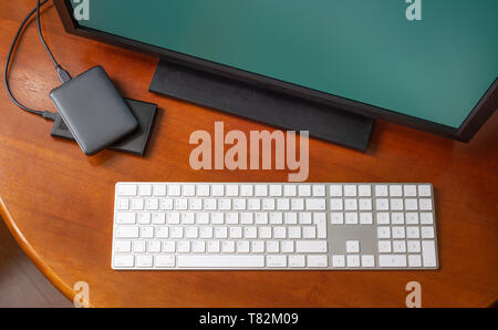 A computer monitor, a digital wireless keyboard and hard disk drives on a round wooden desk Stock Photo
