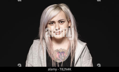 Smiling woman with white hair and tattoo on body looking at camera. Woman with fashion piercing on lips and cheeks isolated on black background. Stock Photo