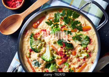 overhead view of  broccoli cheddar cheese soup with green peas and vegetables in a metal casserole on a grey concrete table with wooden spoon, view fr Stock Photo