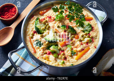 overhead view of  broccoli cheddar cheese soup with green peas and vegetables in a metal pot on a grey concrete table with wooden spoon, view from abo Stock Photo