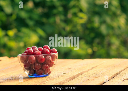 Red ripe cherries in glass bowl on old wooden boards with blurred green background. Stock Photo