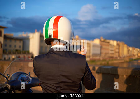 Amazing shot of an Italian man wearing a helmet with the Italian flag standing next to his scooter taking a photo during sunset in Florence, Italy Stock Photo