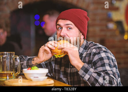 Brutal hipster bearded man sit at bar counter. High calorie food. Cheat meal. Delicious burger concept. Enjoy taste of fresh burger. Hipster hungry man eat burger. Man with beard eat burger menu. Stock Photo