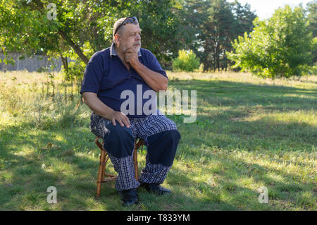 Nice outdoor portrait of Ukrainian thoughtful senior man sitting on a stool in summer garden Stock Photo