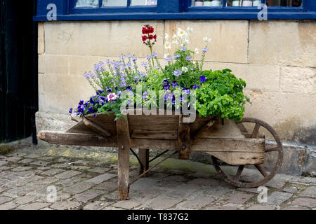 Old wooden wheelbarrow with spring flowers, High Street, Burford, Oxfordshire, England, UK Stock Photo