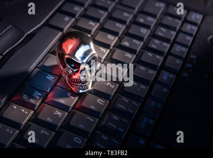 A skull made of shiny metal lies on a keyboard of a computer with strong red reflections Stock Photo