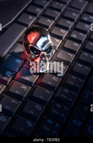 A skull made of shiny metal lies on a keyboard of a computer with strong red reflections Stock Photo
