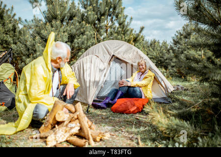 Senior couple in yellow raincoats having fun while making fireplace at the campsite near the tent in the woods Stock Photo