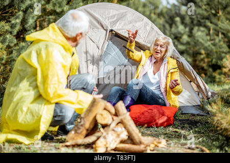 Senior couple in yellow raincoats having fun while making fireplace at the campsite near the tent in the woods Stock Photo