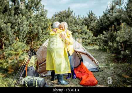 Lovely senior couple in yellow raincoats hugging together while camping with tent in the pine forest Stock Photo