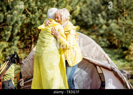 Lovely senior couple in yellow raincoats hugging together while camping with tent in the pine forest Stock Photo