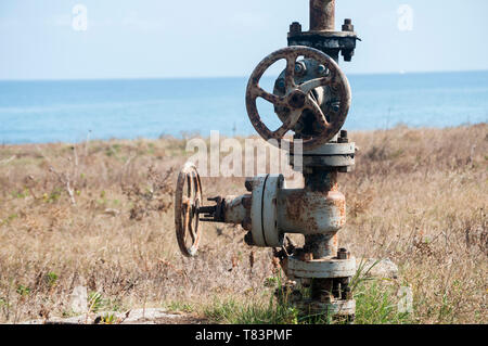 Old rusty weathered aged valve of abandoned pipe system equipment closeup on seashore and blue water background in summer day Stock Photo