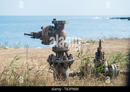 Old rusty weathered aged valve of abandoned pipe system equipment closeup on seashore and blue water background in summer day Stock Photo