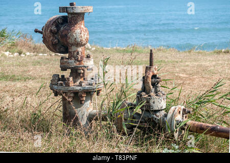 Old rusty weathered aged valve of abandoned pipe system equipment closeup on seashore and blue water background in summer day Stock Photo