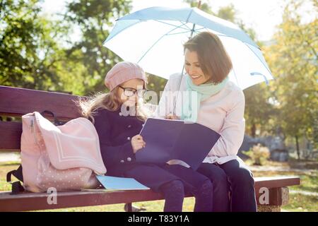 Mom and daughter are resting together on a bench in the city park, the girl in glasses reads to her mother a school notebook, and talks about school l Stock Photo