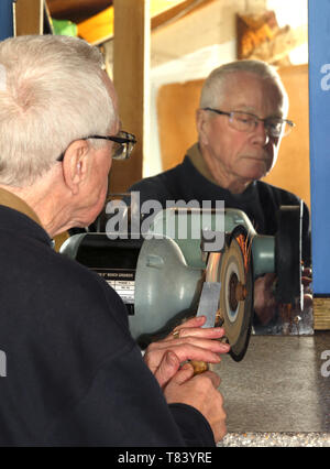 A man in his sixties sharpening a chisel using a bench grinder in a home workshop. Stock Photo