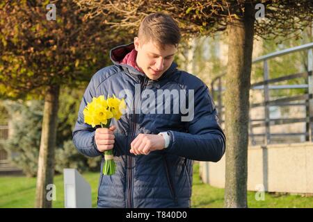 Young man with bouquet of spring flowers looks at wrist watch, urban background, man waiting his girlfriend. Stock Photo
