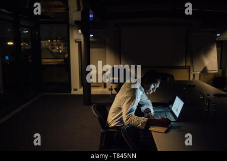 Young businessman in office at night making notes from laptop Stock Photo