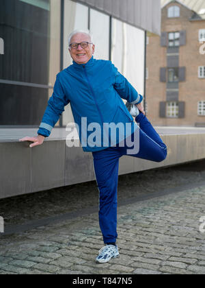 Senior man in tracksuit doing warmup exercises on cobbled street, portrait, Copenhagen, Hovedstaden, Denmark Stock Photo