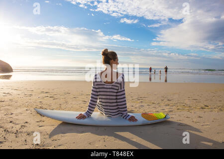 Female surfer sitting on surfboard at beach, rear view, Cape Town, Western Cape, South Africa Stock Photo
