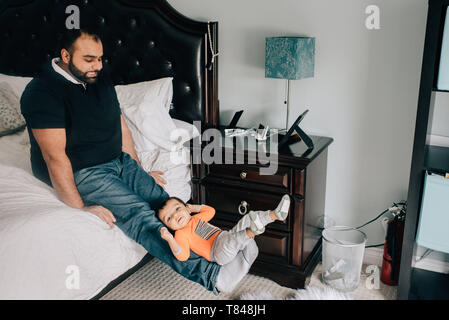 Father sitting on bed with baby son on feet Stock Photo