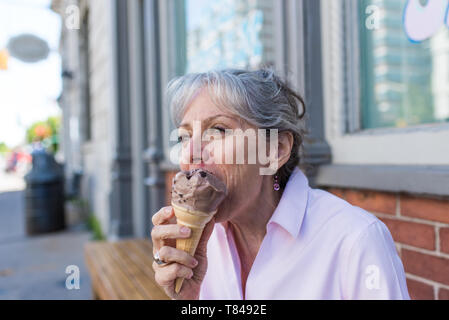 Senior woman sitting on sidewalk eating chocolate ice cream cone Stock Photo