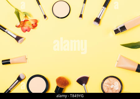 Flat lay frame of women's makeup concealers and brushes on yellow background with empty space in middle. Stock Photo