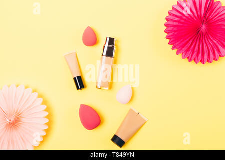 Festive flat lay arrangement makeup products cosmetic foundation next to egg shaped sponge applicators and tissue paper decorations, top view on yello Stock Photo