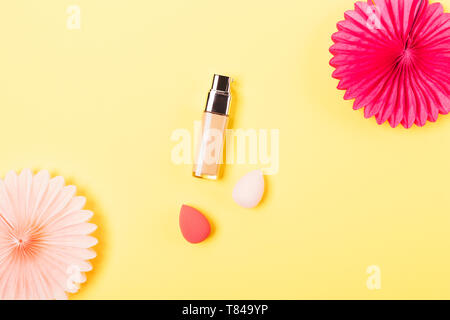 Makeup foundation cream next to egg shaped sponge applicators and festive tissue paper decorations on yellow background, flat lay. Stock Photo