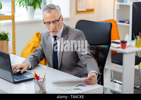 Busy tired grey-haired man in clear glasses reading papers and checking laptop Stock Photo