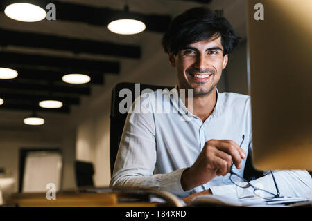Smiling young man working at the office late at night, using computer Stock Photo